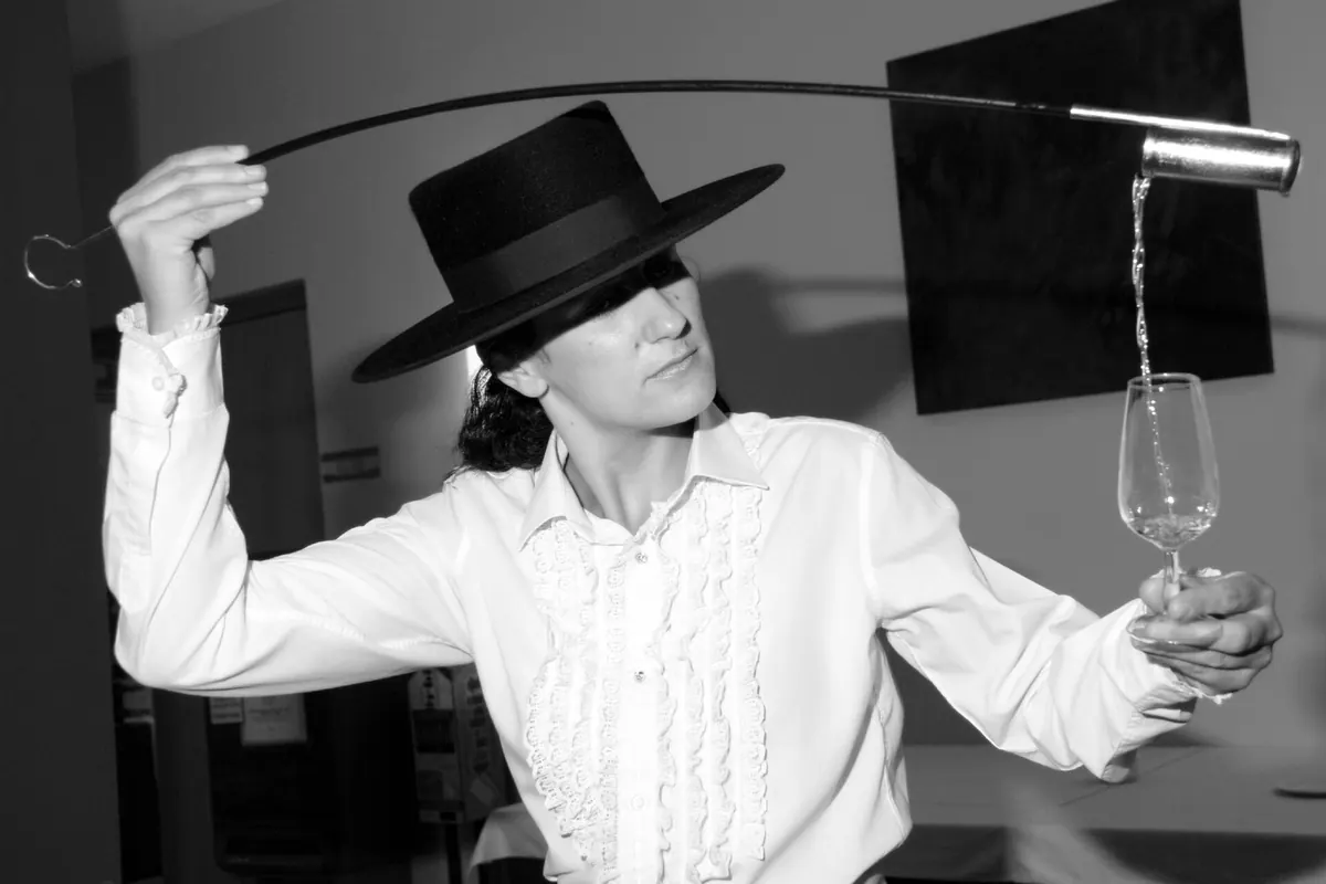 A woman in a wide-brimmed black hat pouring sherry from a long venencia into a glass held at arm's length, in black and white